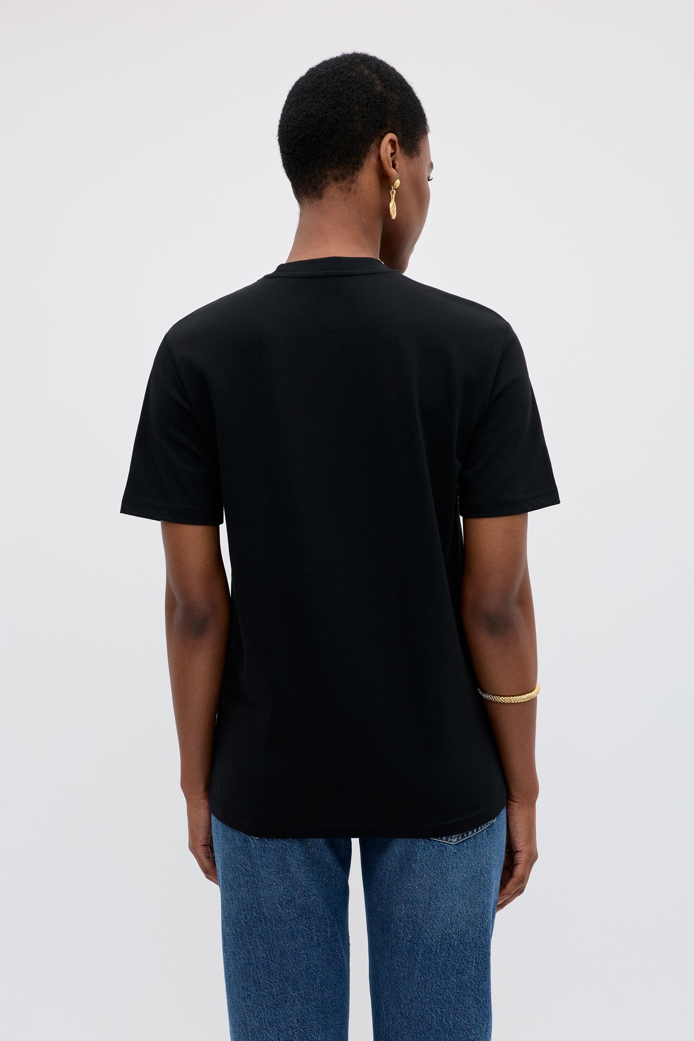 A person with short hair, seen from behind, wears the Charity - Rixo x War Child Black T-shirt, blue jeans, a gold bracelet, and a gold hoop earring against a plain light background.