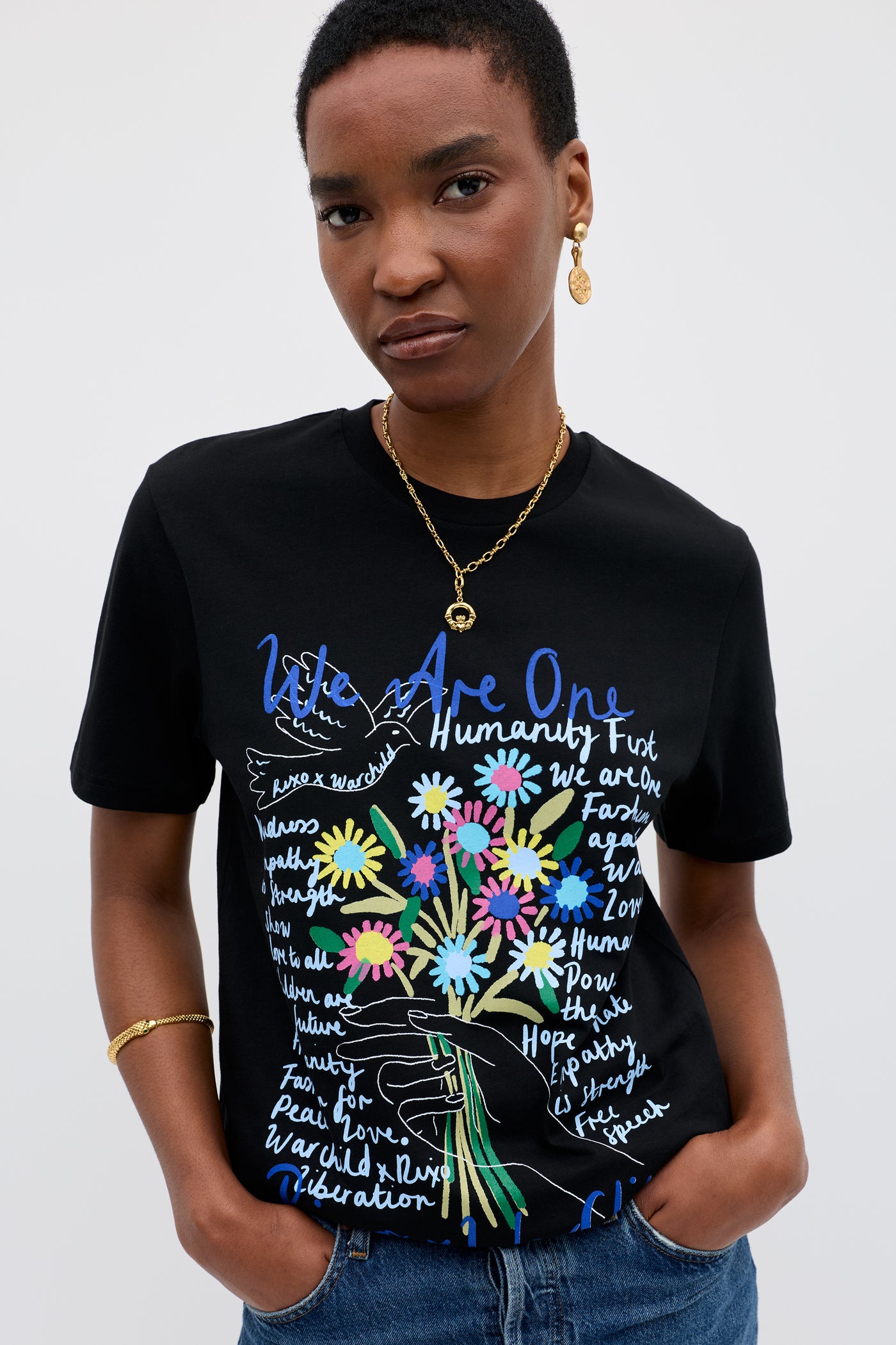 A person with short hair and gold jewelry stands against a plain background, wearing the Charity - Rixo x War Child Black t-shirt. The 60s-inspired tee features colorful flowers and handwritten words like "We Are One" and "Humanity First.