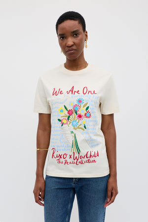 A person stands against a plain background wearing the Charity - Rixo x War Child Cream T-shirt, a 60s-inspired design featuring a colorful bouquet and handwritten "We Are One" text from The Peace Collection.