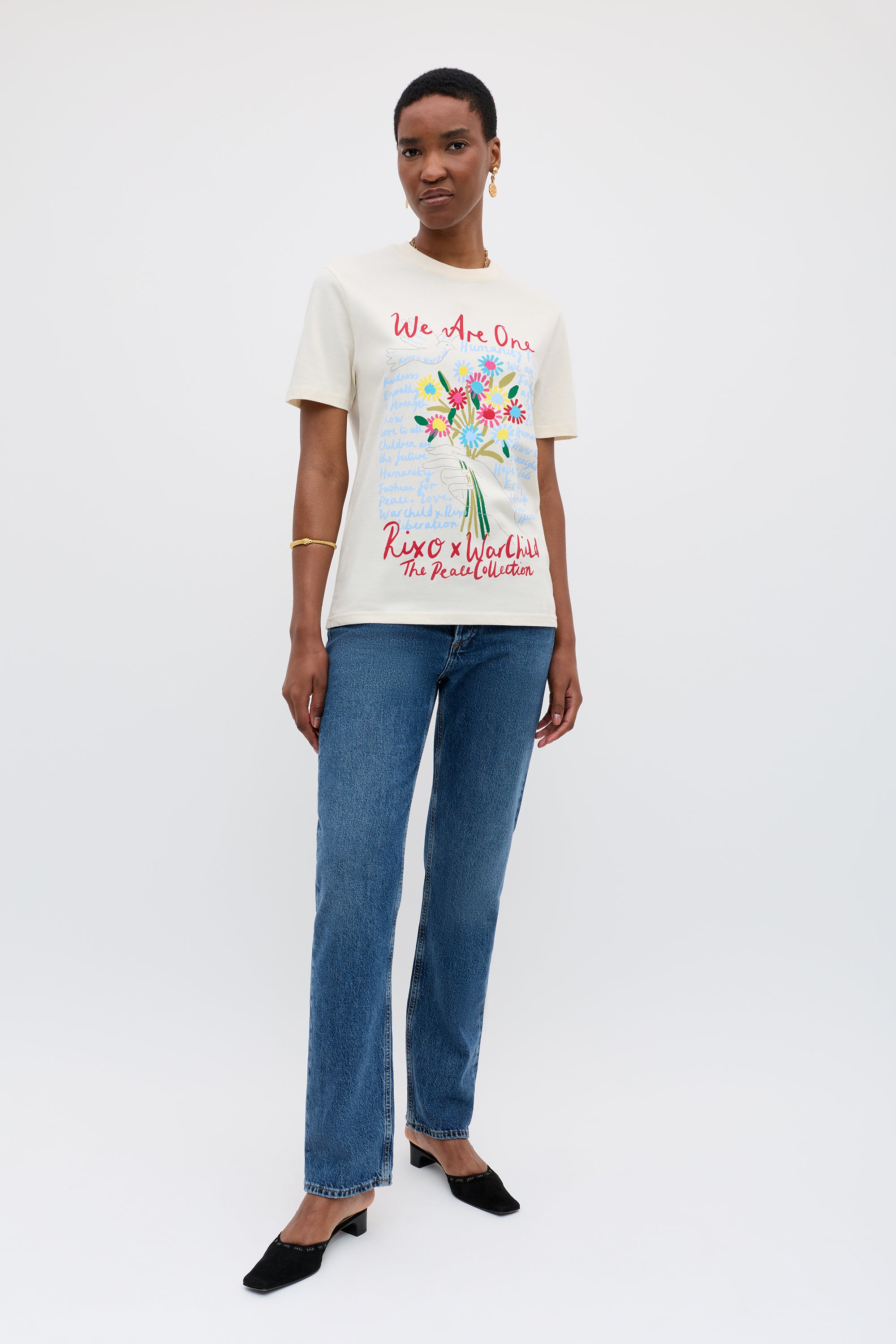 A woman stands against a plain background, wearing the Charity - Rixo x War Child Cream T-shirt featuring a colorful bouquet graphic and text, paired with blue jeans, black heels, and gold jewelry.