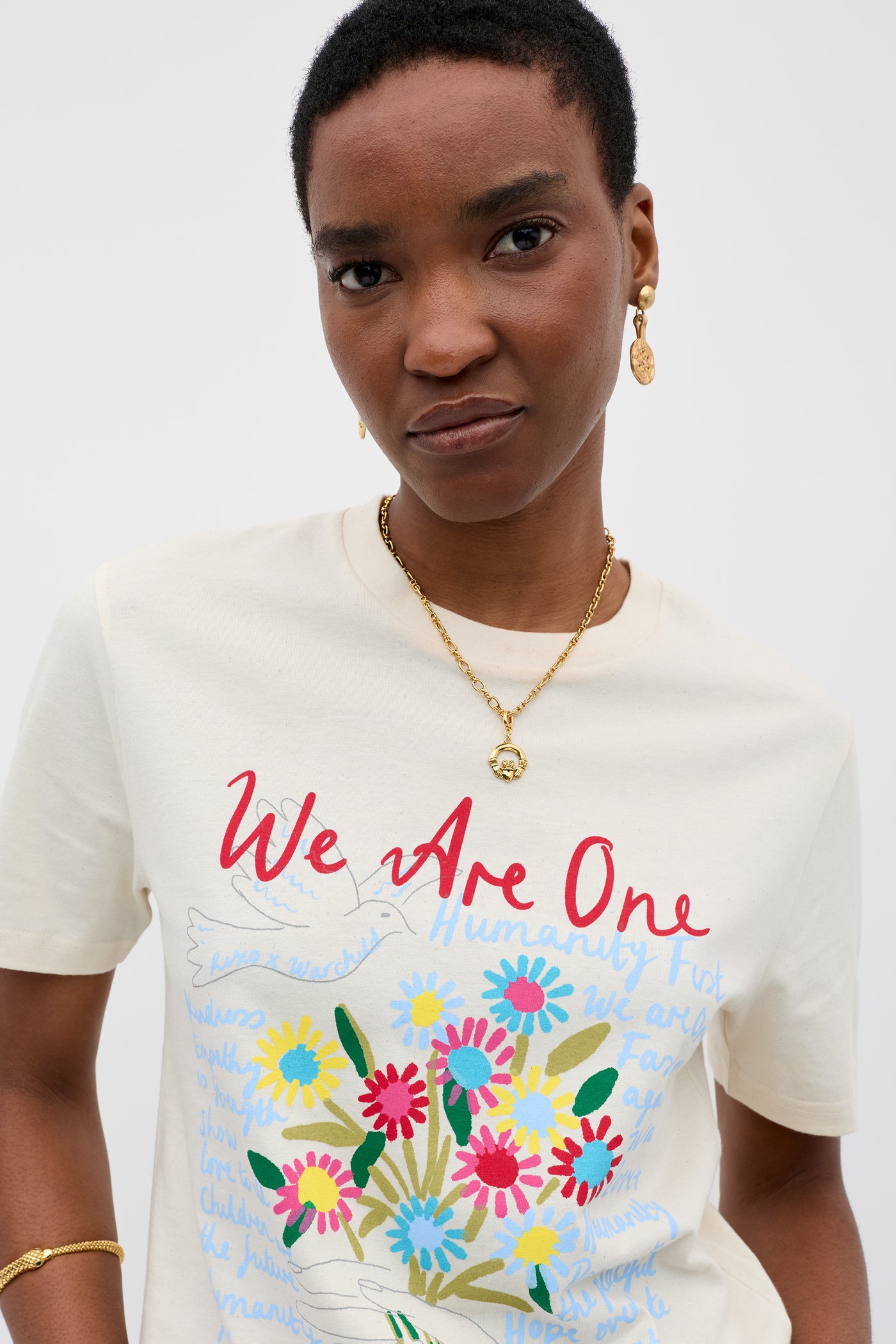 Wearing the Charity - Rixo x War Child Cream T-shirt, a person with short hair and gold jewelry stands against a plain light background; the cream tee features colorful flowers, a white dove, and red "We Are One" text in 60s-inspired style.