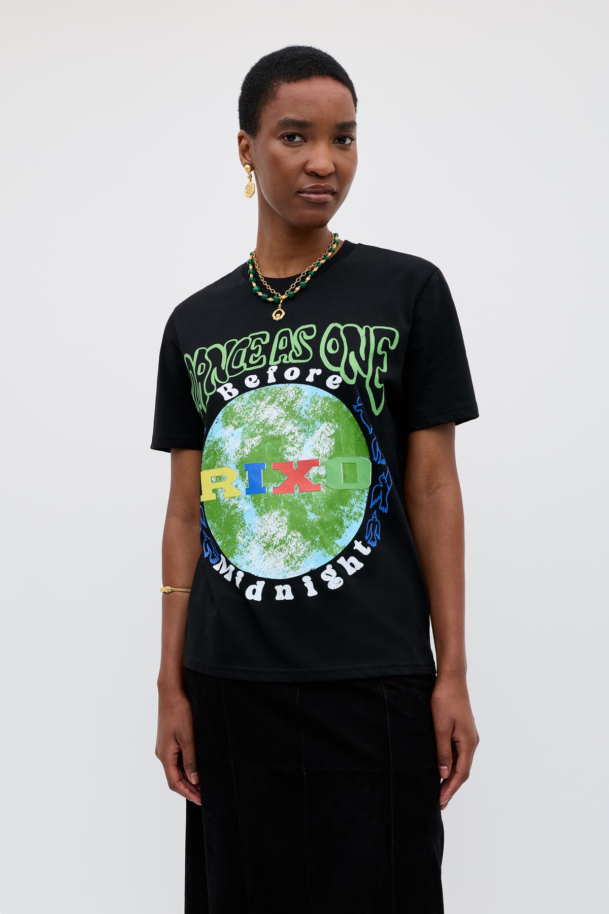 A person with short hair wears the Charity - Rixo x War Child x Before Midnight Black T-shirt with a green and blue earth motif and VOICES AS ONE BEFORE MIDNIGHT text, styled with a necklace, earrings, and black skirt—ideal for the festival.