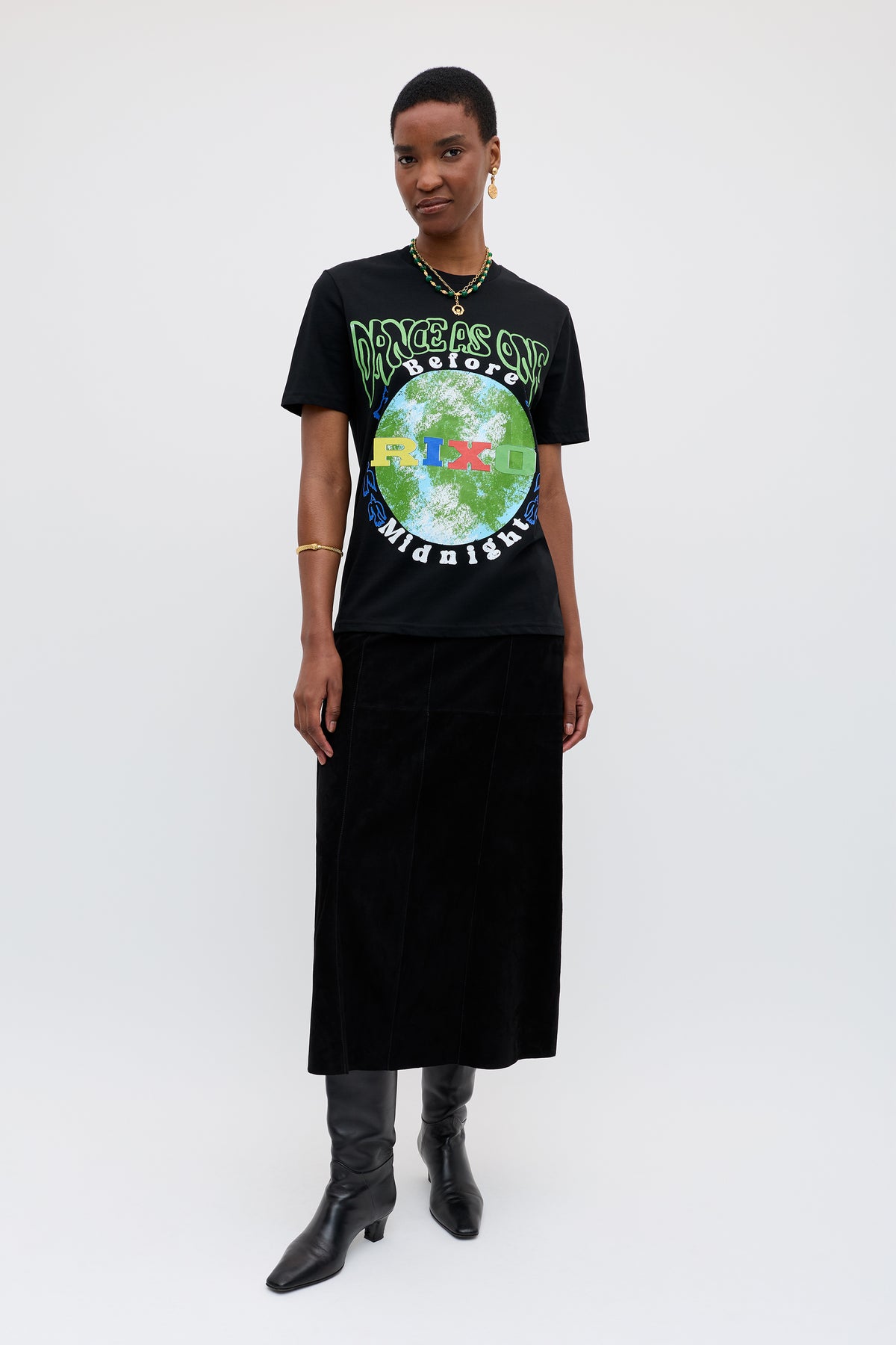 A person stands against a plain background wearing the Charity - Rixo x War Child x Before Midnight Black T-shirt with globe design, a long black skirt, knee-high boots, and gold jewelry—an ideal look for the Before Midnight festival.