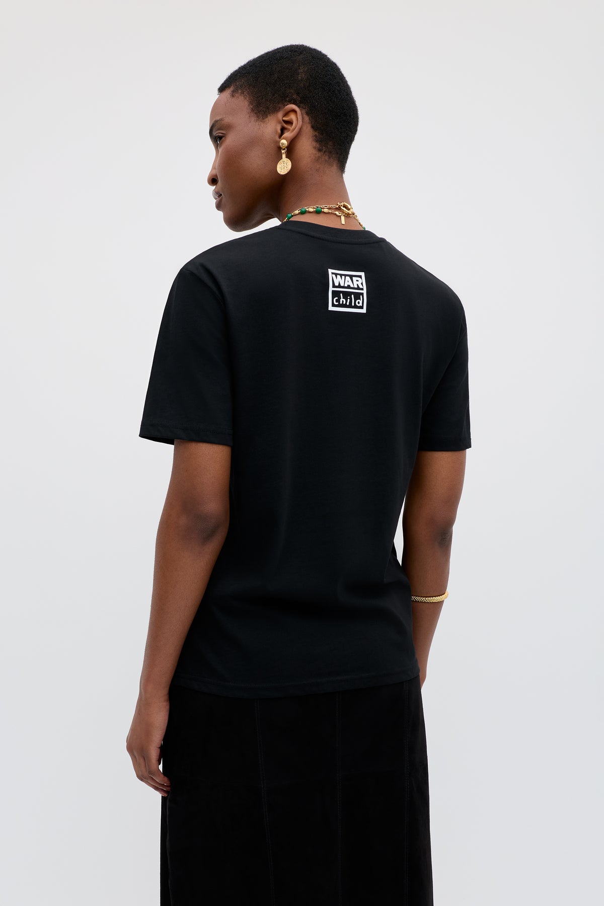 A person with short hair and gold jewelry faces away, displaying the back of the Charity - Rixo x War Child x Before Midnight Black T-shirt. The exclusive tee was designed for DJ Annie Mac’s festival, against a plain light background.