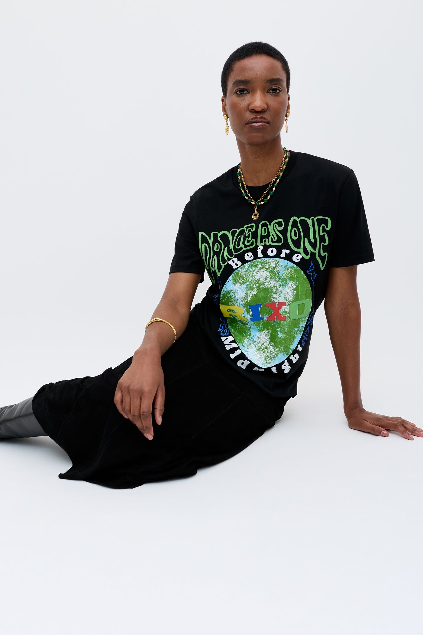 A person with short hair sits against a white background, wearing the Charity - Rixo x War Child x Before Midnight Black T-shirt—featuring green and blue Earth graphics—paired with a long black skirt and boots, ideal for the DJ Annie Mac festival.
