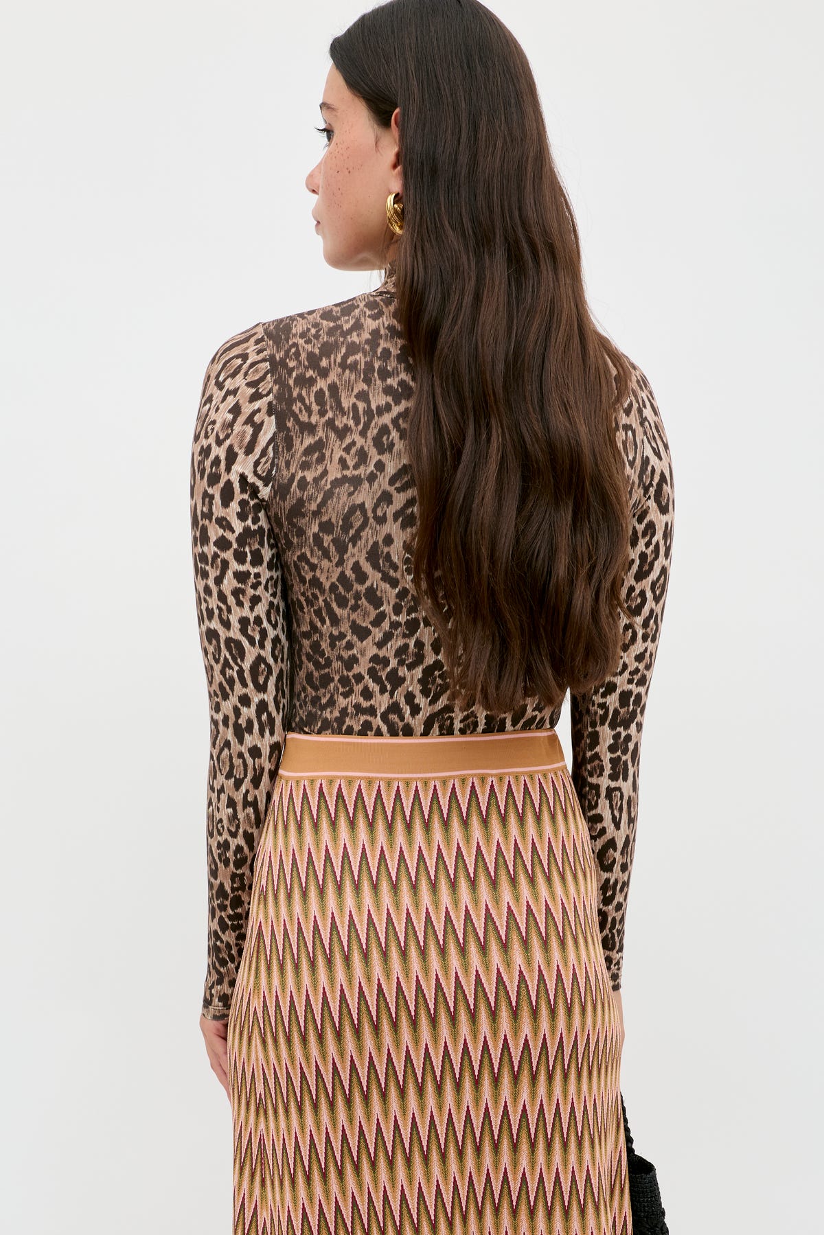 A woman with long brown hair, wearing the Polo - Leopard Brown top and a high-waisted patterned skirt, stands facing away from the camera against a plain light background.