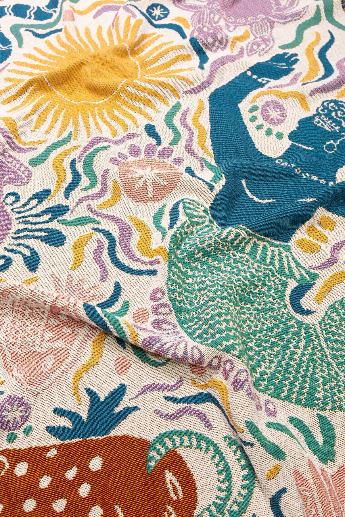 The Athena Throw - Ivory from the Mythos Collection features a colorful cream backdrop with woven texture, illustrated with a green mermaid, blue human figure, and Greek mythology-inspired sea creatures.