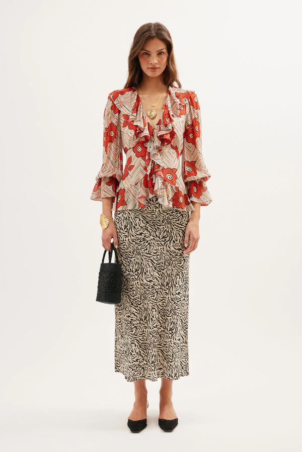 A woman stands against a white background wearing a red floral ruffled blouse, the Ardith - Sun Leo Tan skirt with an elasticated waistband, black pointed shoes, gold jewelry, and holding a small black bag.