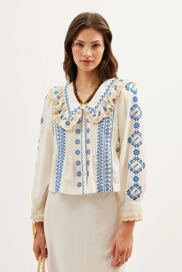 A woman wears the Percy - Mila Embroidery Cream blouse featuring blue embroidery and a ruffled collar, paired with a matching cream skirt. She has long, wavy hair and stands against a plain light background.