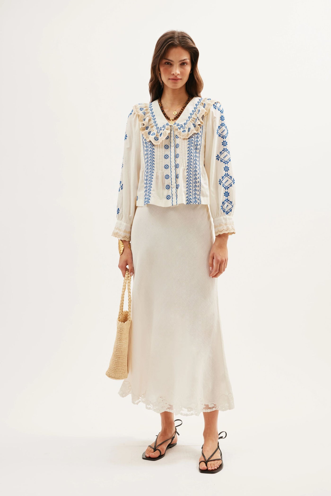 A woman stands against a plain light background wearing the Percy - Mila Embroidery Cream skirt, a vintage-style blue embroidered blouse with a ruffled collar, black strappy sandals, and holding a woven beige bag.