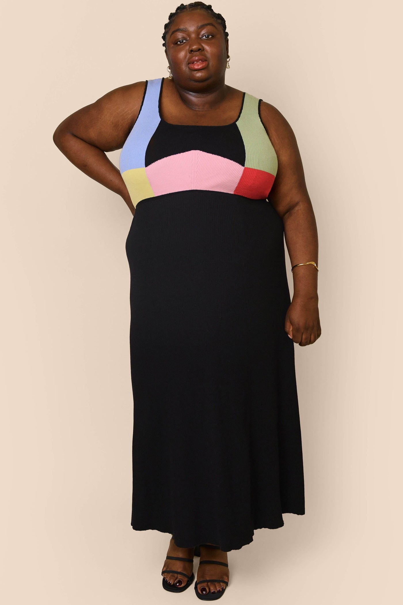 A woman stands against a beige background wearing the Abisola - Geo Multi knit midi dress, showcasing a sleeveless silhouette and vibrant geometric print. She completes the look with braided hair, black sandals, and a gold bracelet.