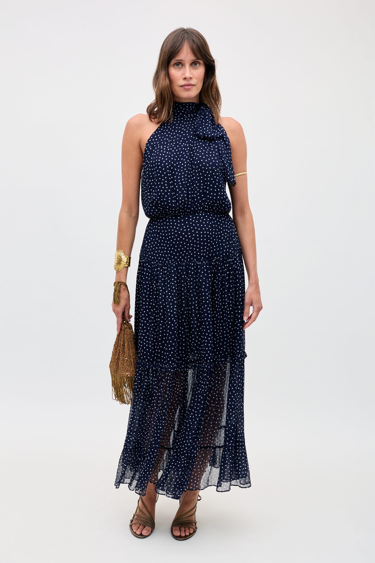 A woman stands against a plain background, wearing the Abi - Mini Navy Polka Dot dress with white polka dots. She accessorizes with a gold fringe handbag, gold jewelry, and brown strappy sandals.