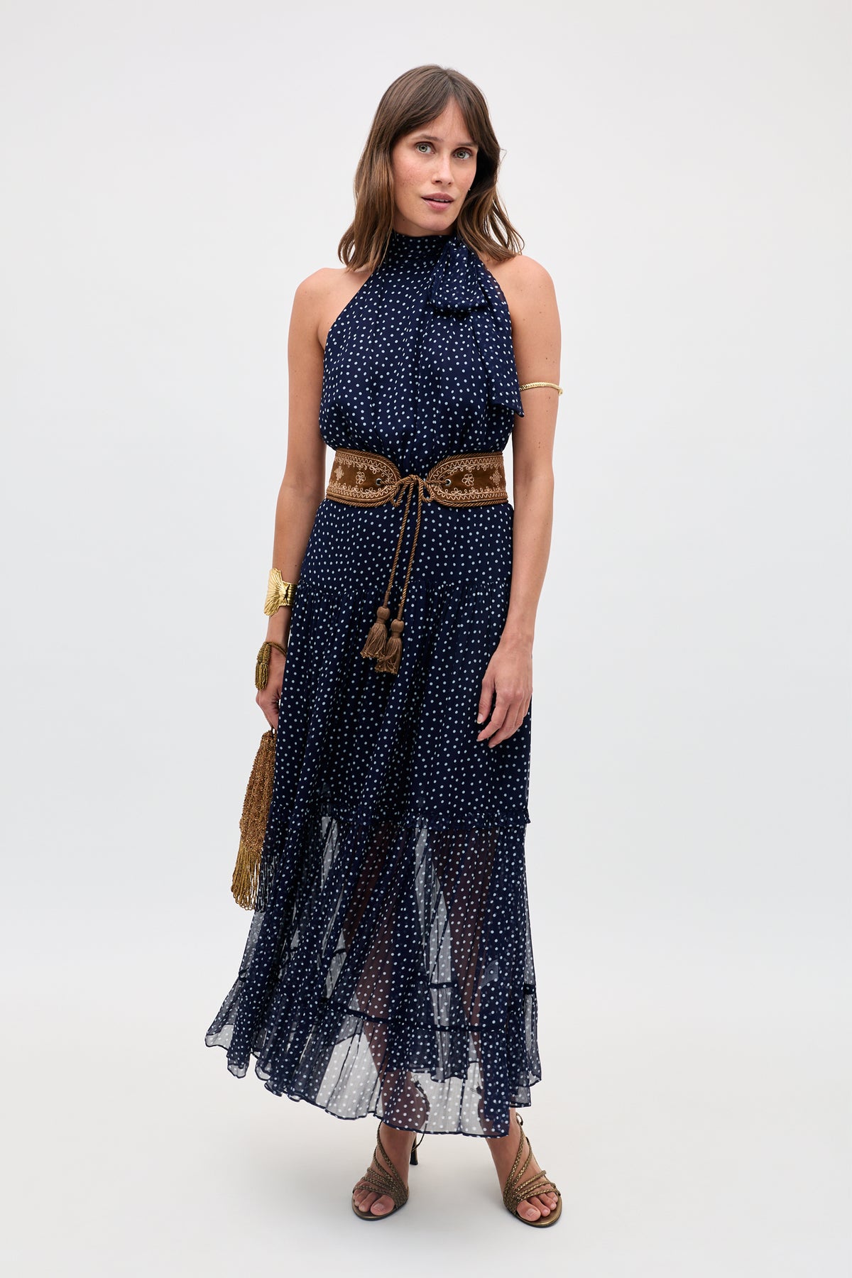 A woman poses against a plain backdrop wearing the Abi - Mini Navy Polka Dot dress with a halter neckline, styled with a tan belt, gold arm cuff, bracelets, brown sandals, and a straw handbag.