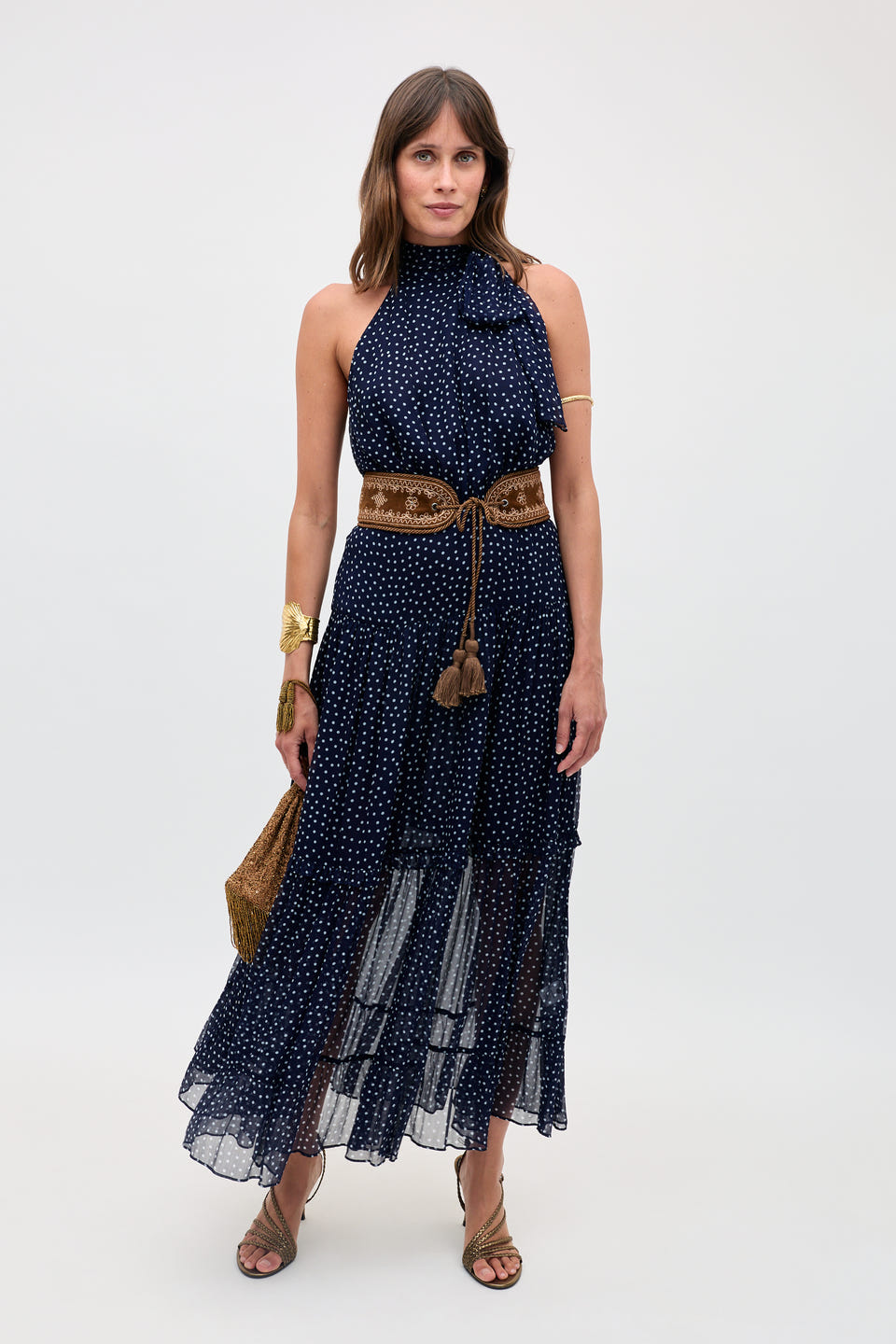 A woman in the Abi - Mini Navy Polka Dot dress, styled sleeveless with a high neckline, accessorized with a brown belt, gold jewelry, tan sandals, and a woven bag, poses against a plain light background.