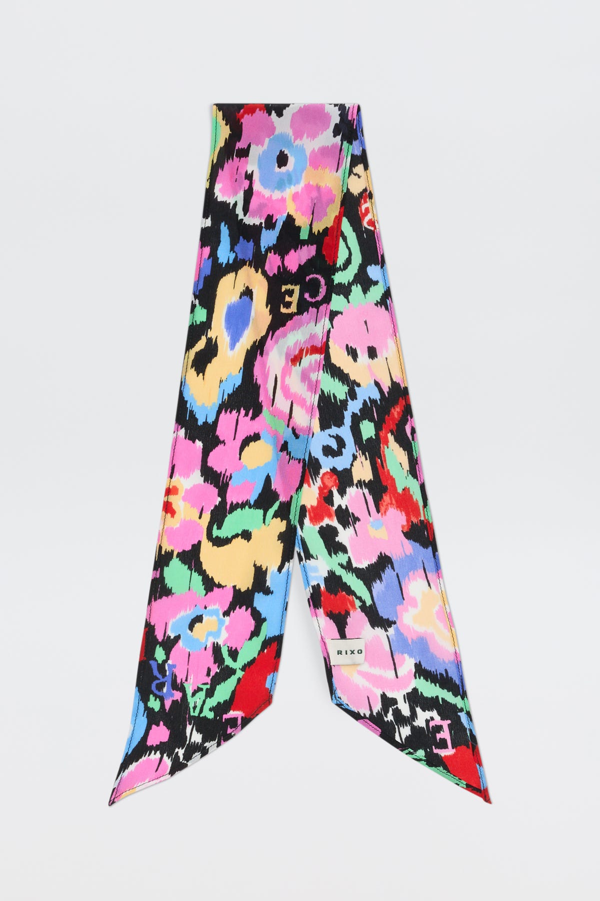 The Alexa - Ikat Bloom Charcoal is a silk scarf with abstract floral patterns in pink, yellow, blue, green, and red on a black background. It features pointed ends, a small brand label, and is displayed against a white backdrop.