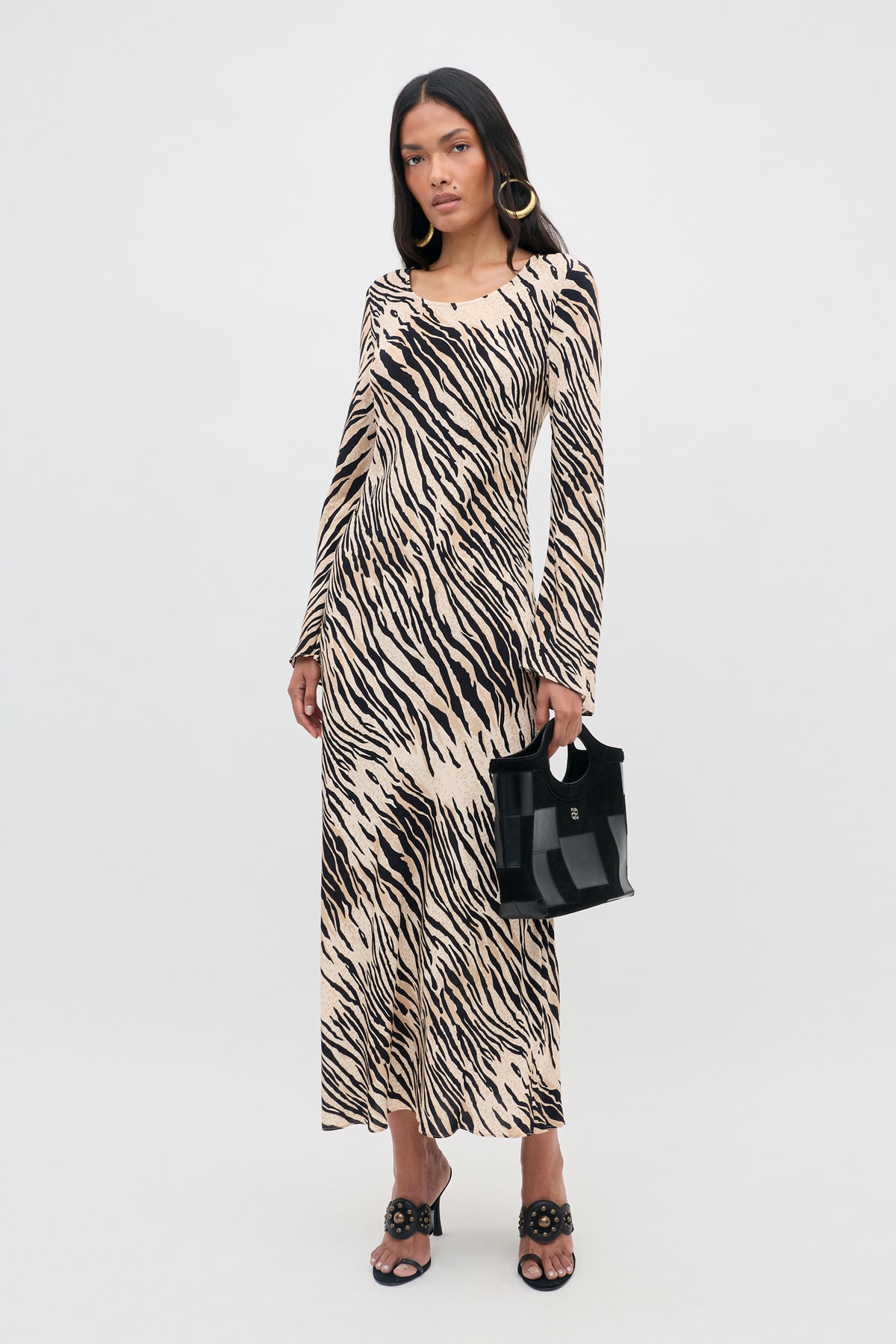 A woman wearing the Amal - Cooper Tiger Beige long-sleeved maxi dress, paired with black sandals and large hoop earrings, holds a small black handbag while standing against a plain light gray background.