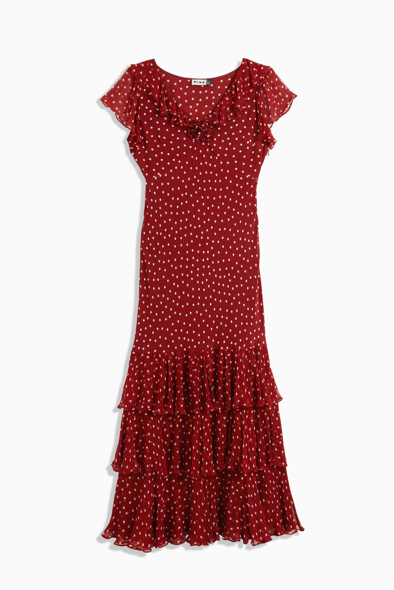 The Amelle Layla Dot Red is a classic ankle-length silk dress in red with white polka dots, featuring short sleeves, a ruffled neckline, and a layered ruffle hem.
