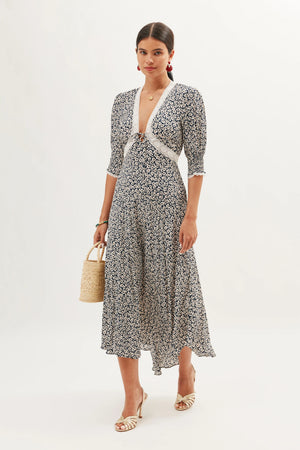 A woman stands against a white background wearing the Amina - English Daisy Deep Navy dress, a navy and white floral midi with a deep V-neckline and lace trim. She accessorizes with a woven handbag, white heeled sandals, and earrings.