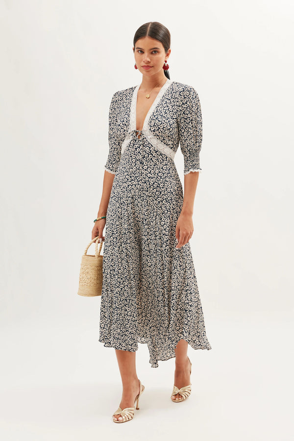 A woman stands against a white background wearing the Amina - English Daisy Deep Navy dress, a navy and white floral midi with a deep V-neckline and lace trim. She accessorizes with a woven handbag, white heeled sandals, and earrings.