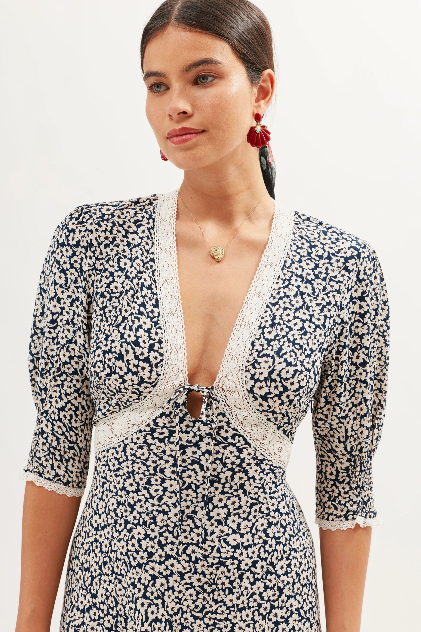 A woman wears the Amina - English Daisy Deep Navy dress, a blue and white floral piece with a lace-trimmed deep V-neck, puffed half sleeves, and keyhole neckline. Her dark hair is pulled back and she accessorizes with red statement earrings.