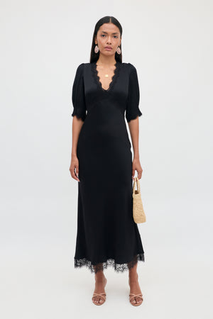 Clad in the Annina - Black dress, she showcases its v-neckline and short sleeves, accented by delicate lace trims at the hem. The bias cut of this ankle-length midi dress flatters her silhouette as she holds a small woven bag. Completing her look with sandals, statement earrings, and layered necklaces, she stands against a plain white background.