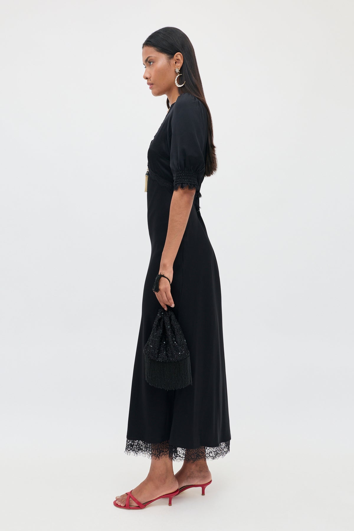 A woman in the Annina - Black bias cut midi dress with delicate lace trims holds a small black bag. She pairs the look with red heeled sandals and large hoop earrings, her straight dark hair worn loose, standing in profile against a plain white background.