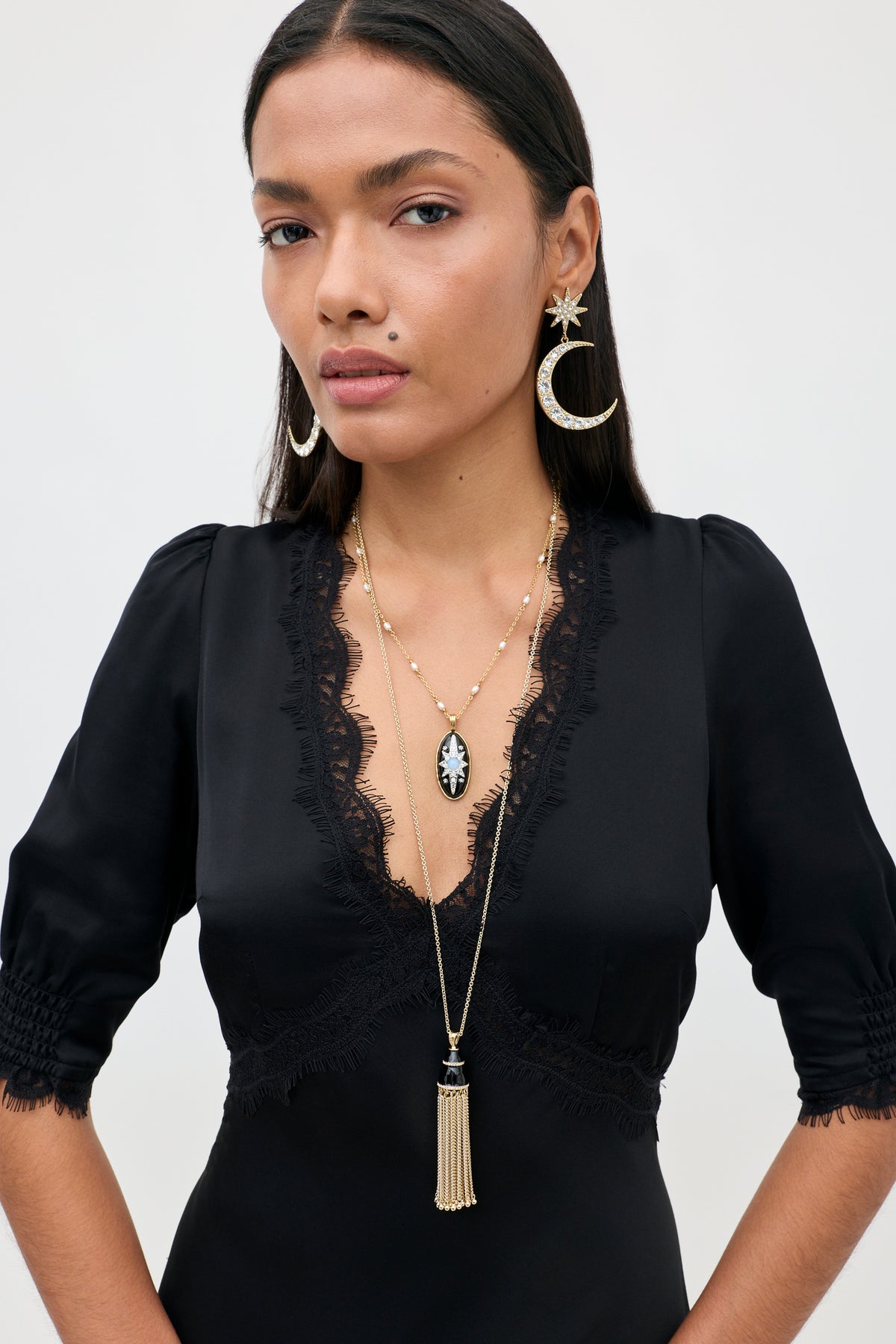 A woman with long dark hair wears the Annina - Black midi dress, which features a deep V-neckline, a bias-cut silhouette, and delicate lace trims. She pairs the dress with gold crescent moon earrings and layered gold necklaces with pendant and tassel details, standing against a plain light background.