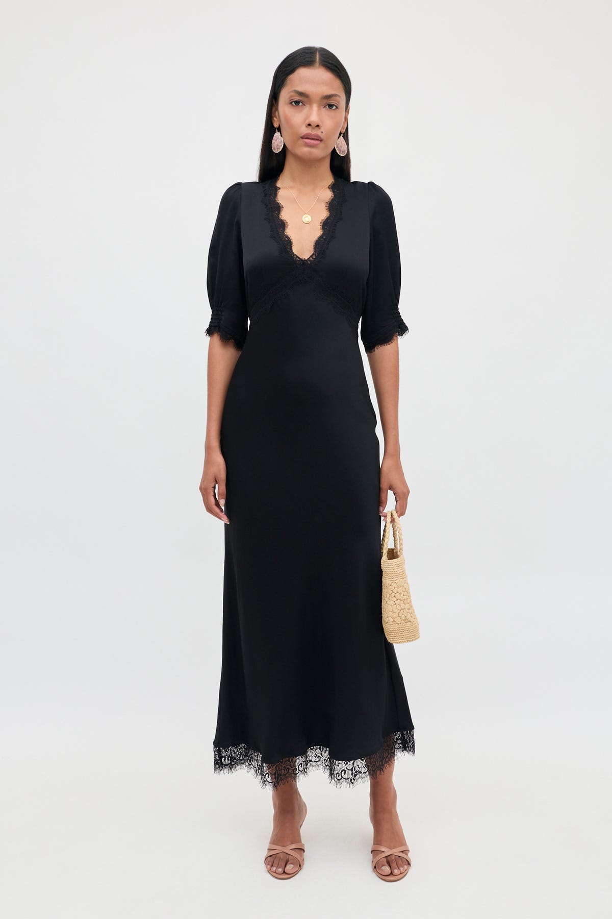 A woman wears the Annina - Black midi dress, featuring a V-neck, short sleeves, delicate lace trims, and an elegant bias cut. She pairs it with brown sandals, gold jewelry, and a small woven bag while standing against a plain light background.