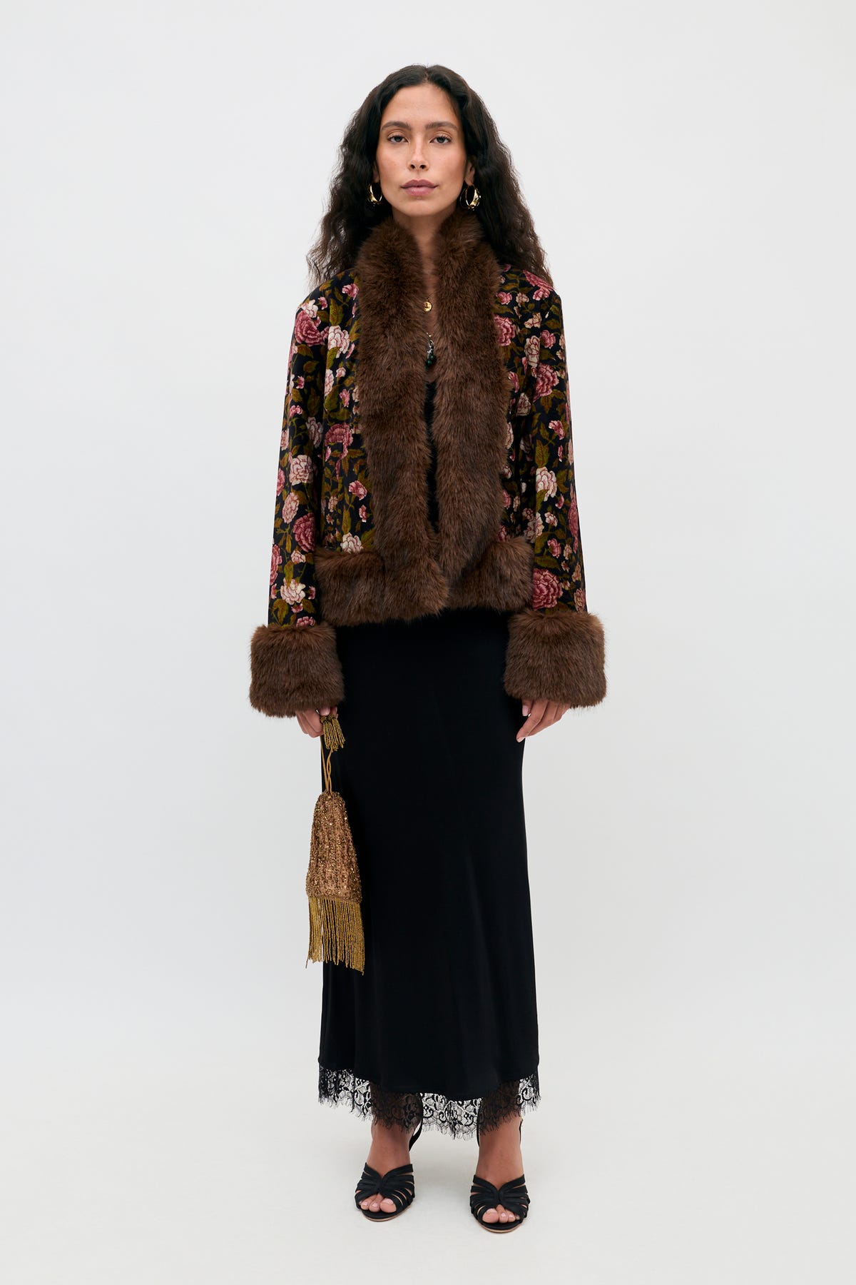 A woman stands against a white background wearing the Annina - Black midi dress, which features delicate lace trims at the hem and a flattering bias cut. She pairs it with a floral jacket with brown fur trim, black sandals, and holds a gold fringed purse.