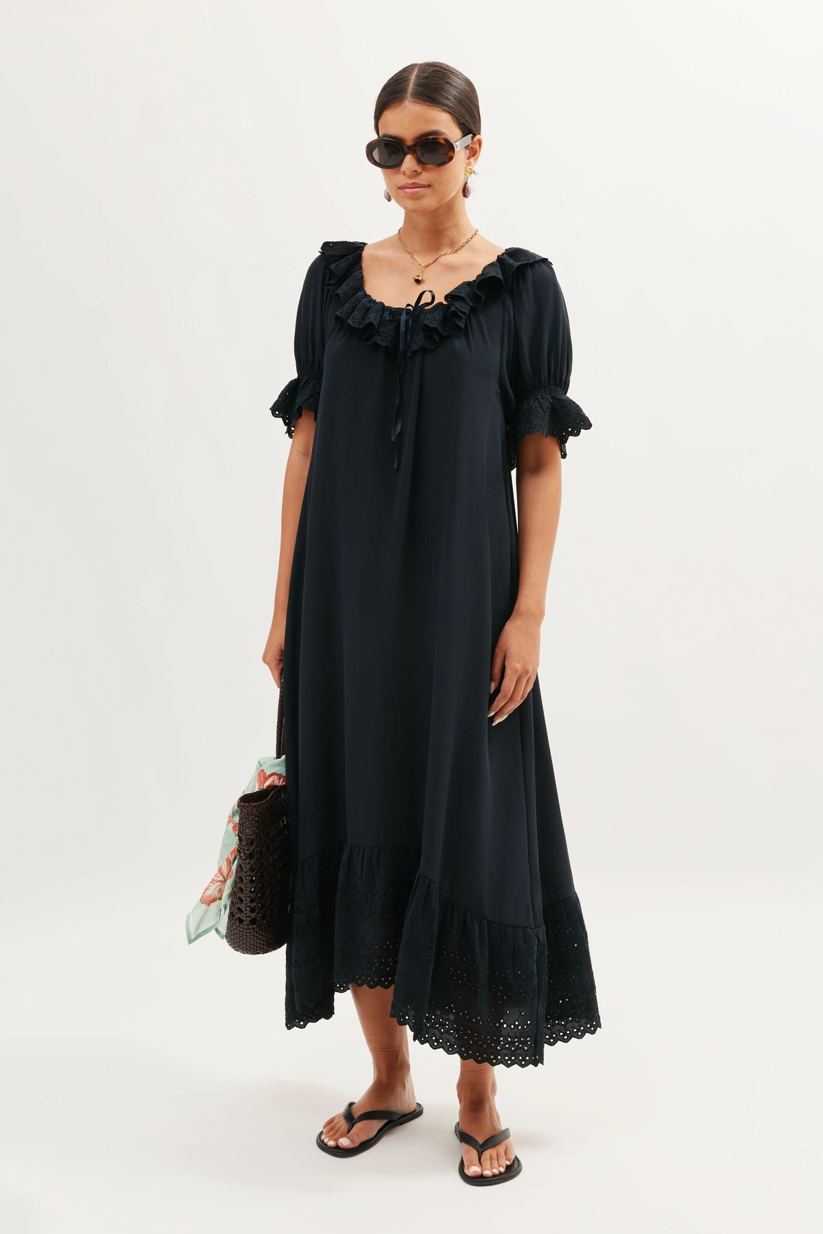 A woman stands against a plain background in the Aprilia Gracie Embroidery Black dress—loose-fitting silk with short ruffled sleeves and lace trim. She pairs it with sunglasses, sandals, a necklace, and a black woven bag accented by a scarf.
