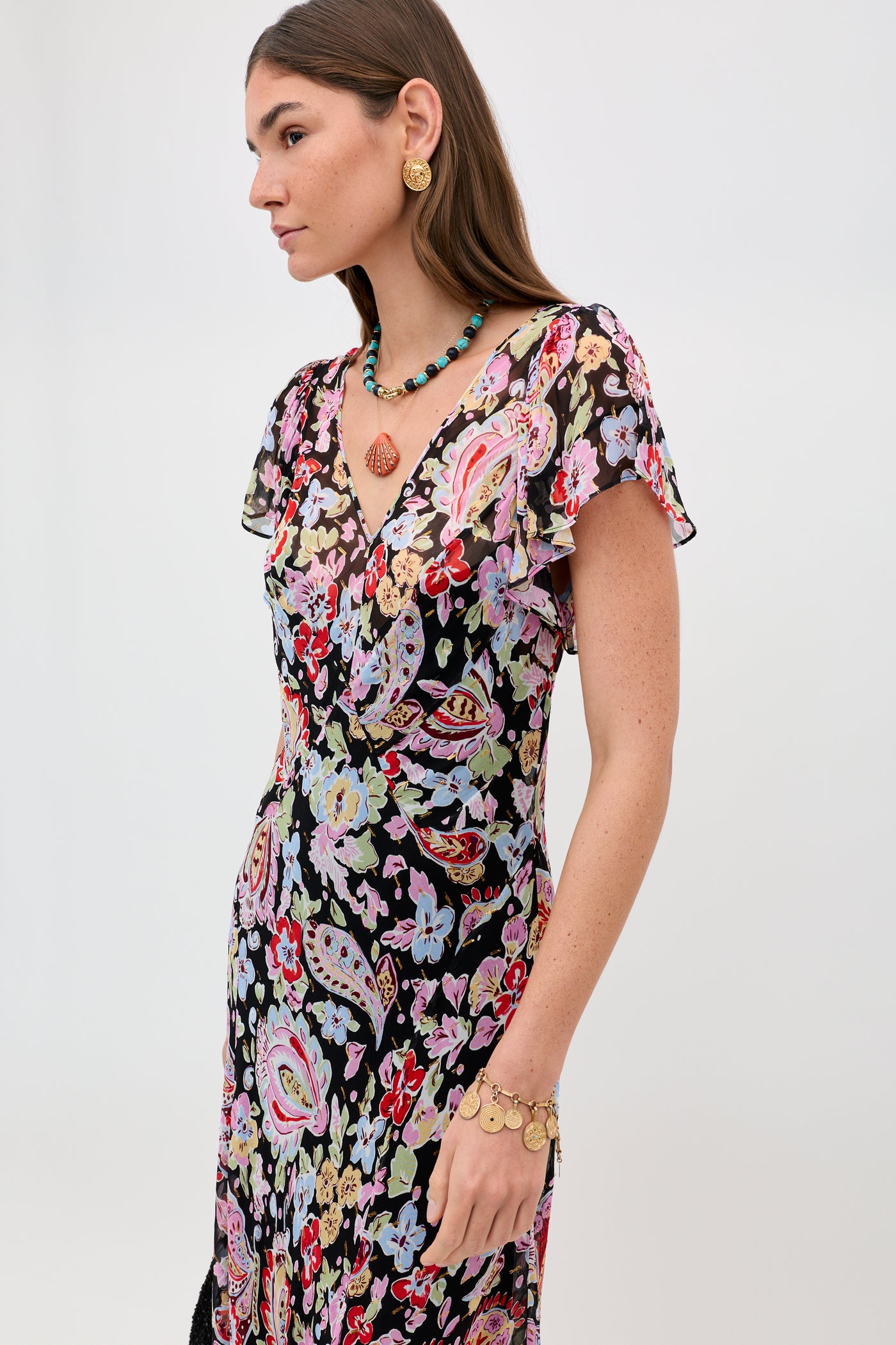 A woman in the Bexley - Cara Floral Black dress, gold earrings, a turquoise beaded necklace, and a gold bracelet stands against a plain light background, looking to the side.