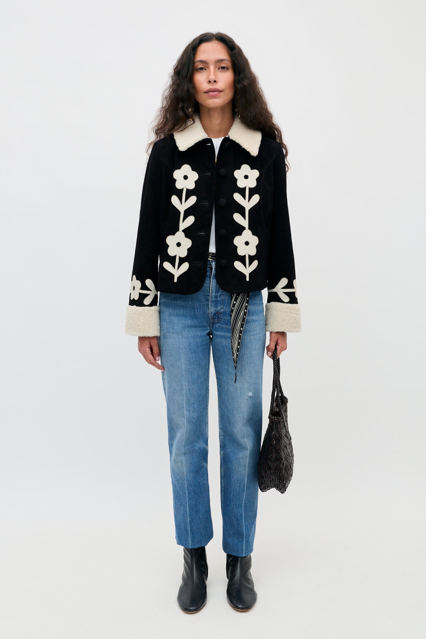 A woman stands facing forward, wearing the Brodie - Retro Daisy Applique Embroidery Black jacket, paired with blue jeans, black boots, and holding a woven bag in her left hand.