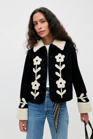 A woman with long wavy hair wears the Brodie - Retro Daisy Applique Embroidery Black jacket, featuring white floral patterns and cream fur cuffs and collar, paired with blue jeans and holding a bag. She stands against a plain light background.