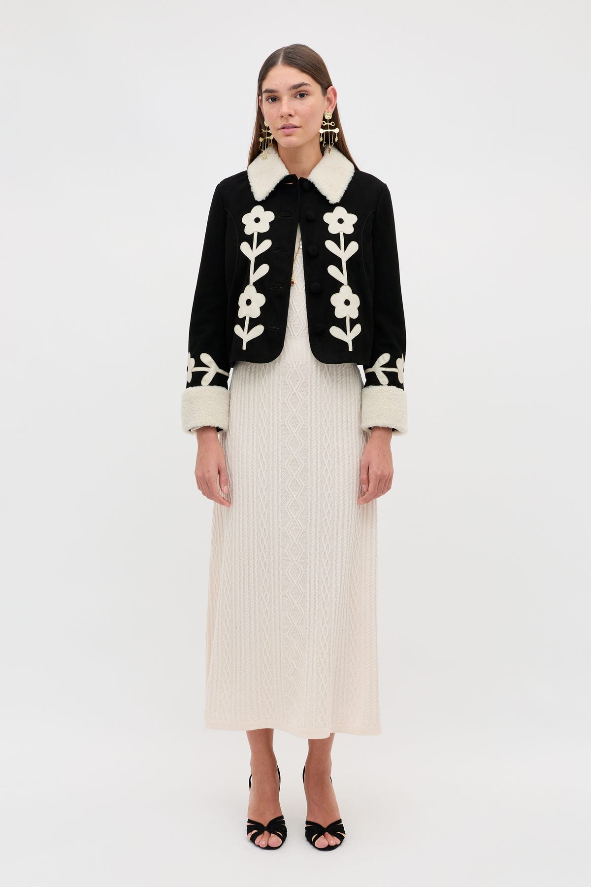 A woman wearing the Brodie - Retro Daisy Applique Embroidery Black jacket over a cream knit dress stands against a plain white background. She pairs the look with statement earrings, black open-toe heels, and has long straight hair.