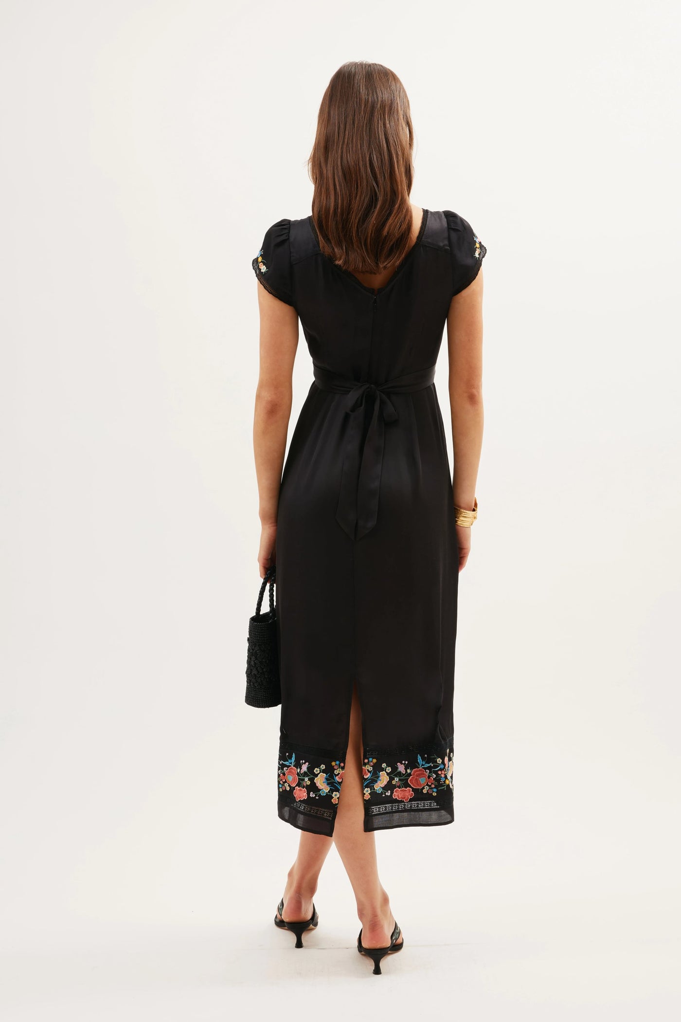 Seen from behind, a woman wears the Carey - Orchard Embroidery Black dress, featuring feminine embroidery at the hem, short sleeves, a waist tie, and a back slit. She holds a black handbag and wears open-toe heels.