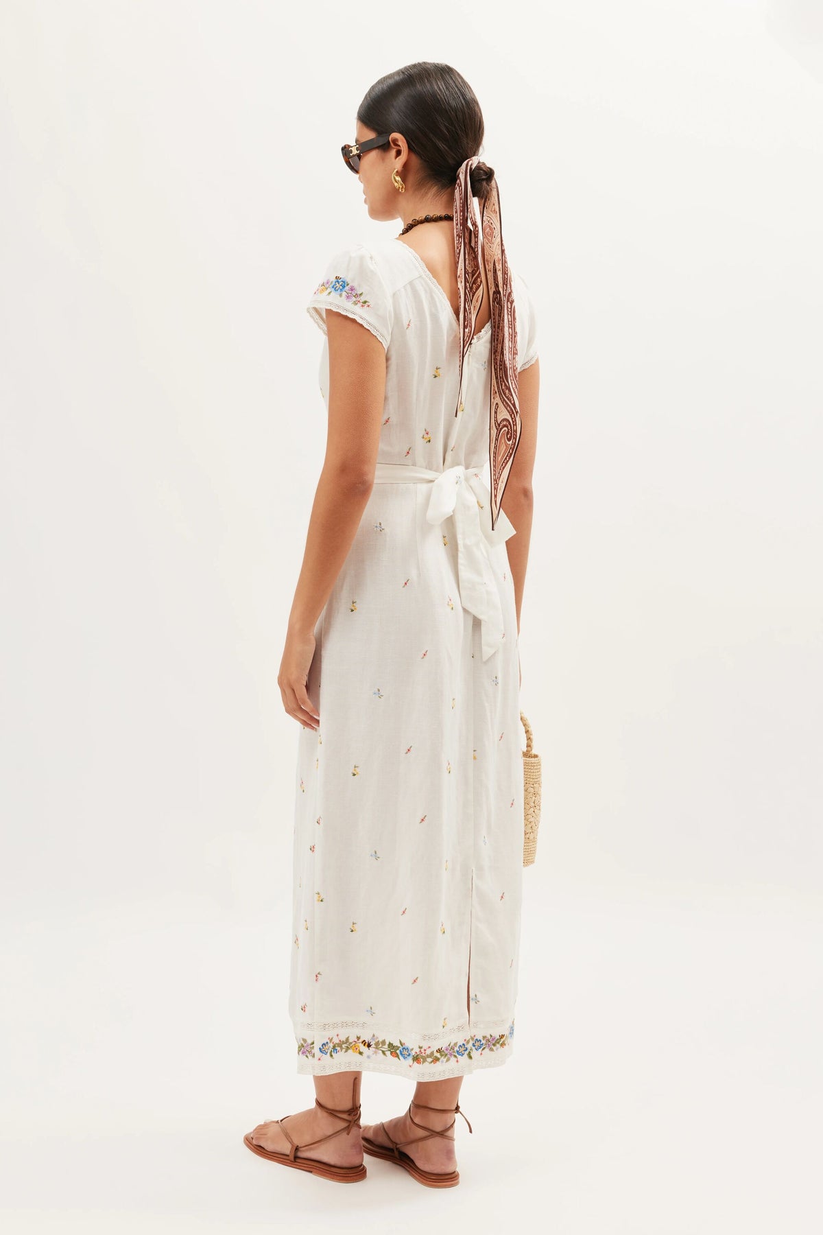 A woman with dark hair in a low bun, patterned scarf, sunglasses, brown sandals, and a straw bag is shown from the back wearing the Carey - Meadow Embroidery Ivory dress against a plain background.