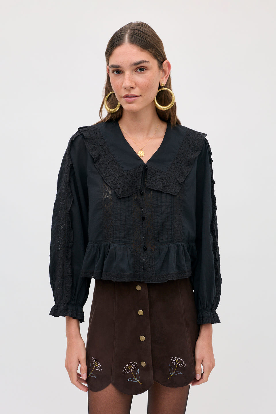 A woman wears a Caris - Shasta Daisy Embroidery Chocolate skirt with small floral embroidery along the scalloped hem, paired with a black long-sleeved blouse featuring a lace-trimmed collar.