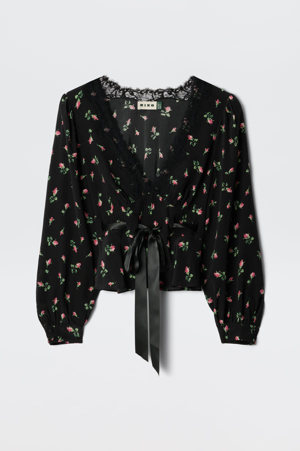 The Charis - Sienna Bud Black blouse by RIXO features black fabric with long sleeves, a delicate pink and green floral print, lace-trimmed deep V-neckline, and a wide satin ribbon bow at the waist for romantic femininity.