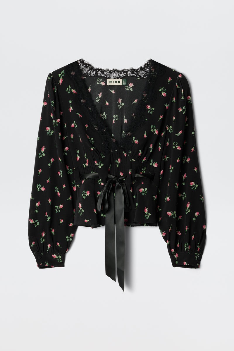 The Charis - Sienna Bud Black blouse by RIXO features black fabric with long sleeves, a delicate pink and green floral print, lace-trimmed deep V-neckline, and a wide satin ribbon bow at the waist for romantic femininity.