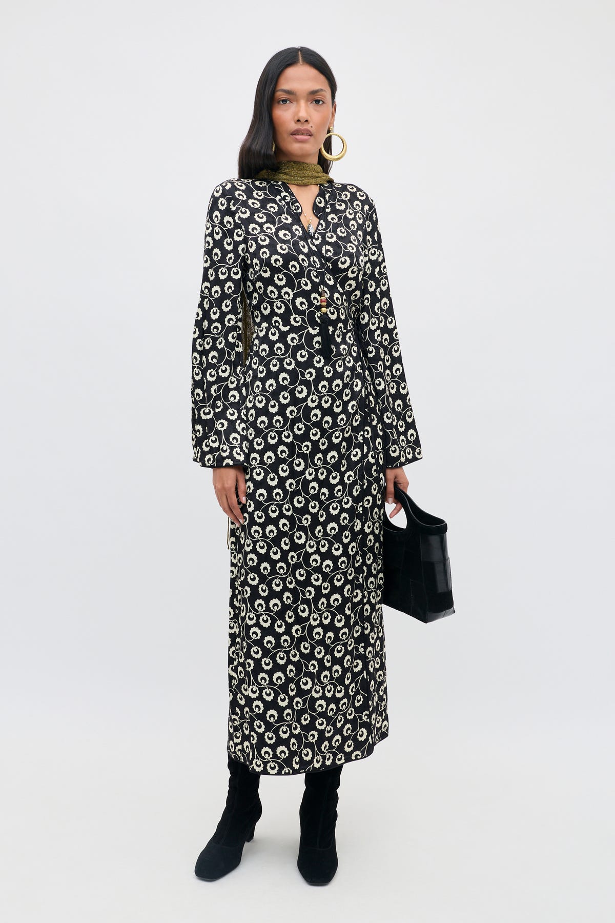 A woman stands against a plain background, wearing the Cindy - Wishing Floral Black long-sleeve patterned dress, paired with black boots, large hoop earrings, a green scarf, and holding a black handbag.