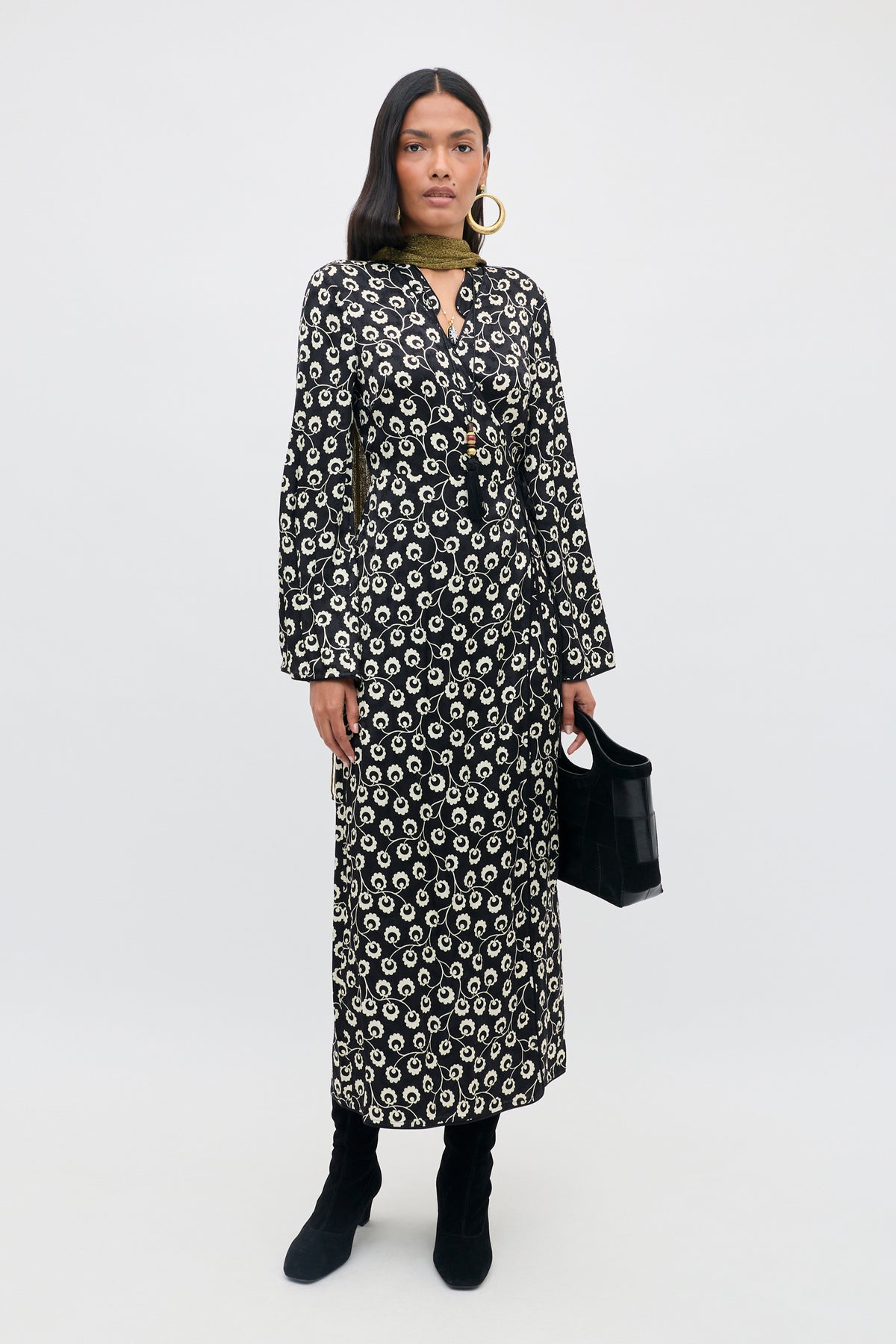 A woman stands against a plain background, wearing the Cindy - Wishing Floral Black long-sleeve patterned dress, paired with black boots, large hoop earrings, a green scarf, and holding a black handbag.