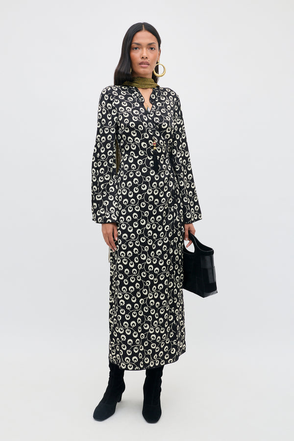 A woman stands against a plain background, wearing the Cindy - Wishing Floral Black long-sleeve patterned dress, paired with black boots, large hoop earrings, a green scarf, and holding a black handbag.