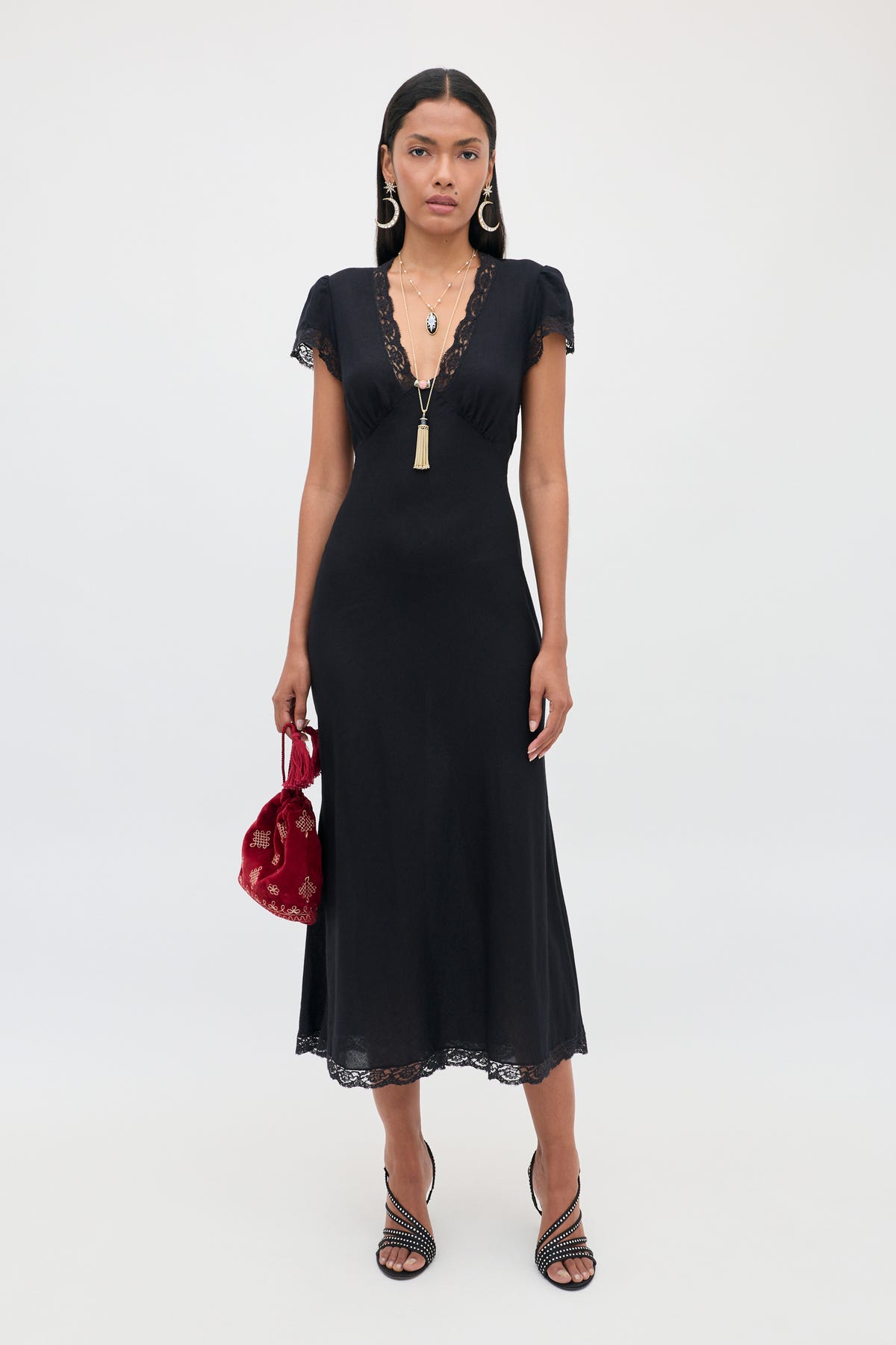 Wearing the Clarice - Black dress, a woman stands against a plain light background, styled with necklaces, black heeled sandals, and a red handbag.