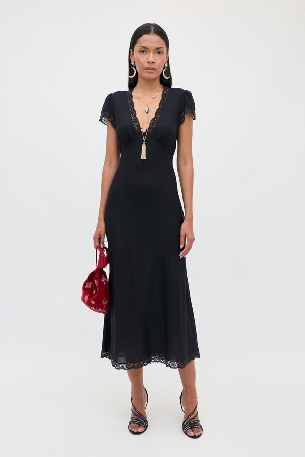 Wearing the Clarice - Black dress, a woman stands against a plain light background, styled with necklaces, black heeled sandals, and a red handbag.