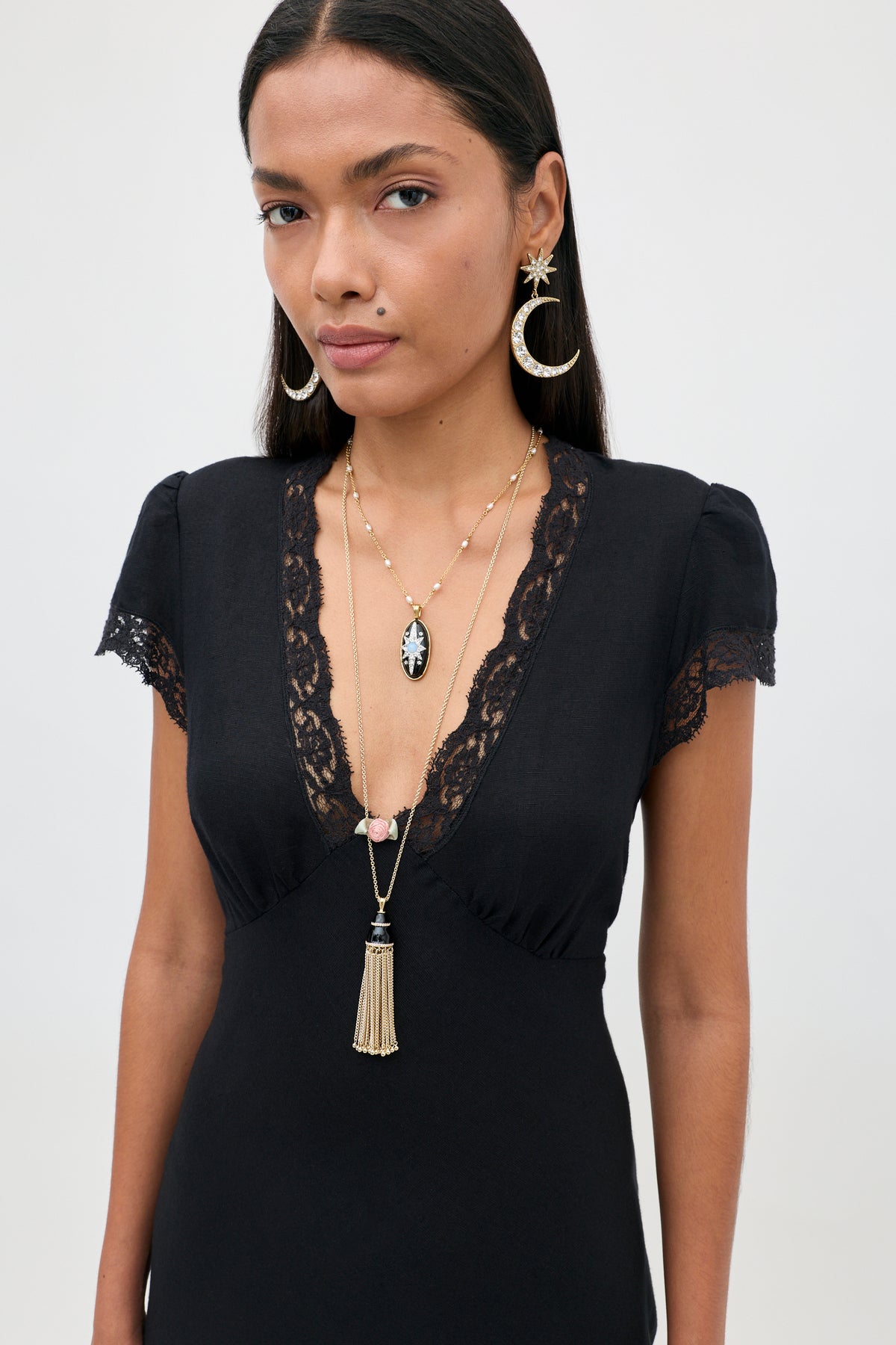 A woman with long dark hair wears the Clarice - Black vintage nightdress with lace trim, layered necklaces, and gold crescent moon earrings, standing confidently against a plain light background and looking at the camera.