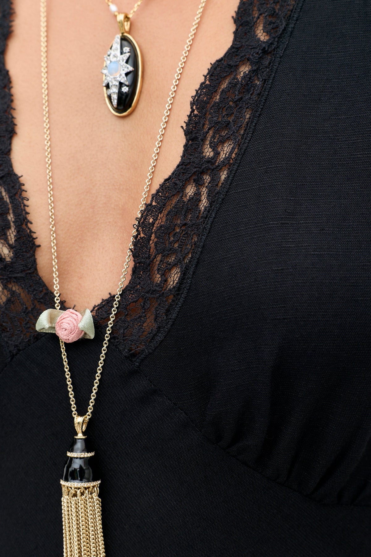 A close-up of a person wearing the Clarice - Black top, featuring vintage-inspired lace trim, along with two necklaces: one adorned with a black oval pendant and star, and the other with a small pink rose and gold tassel.