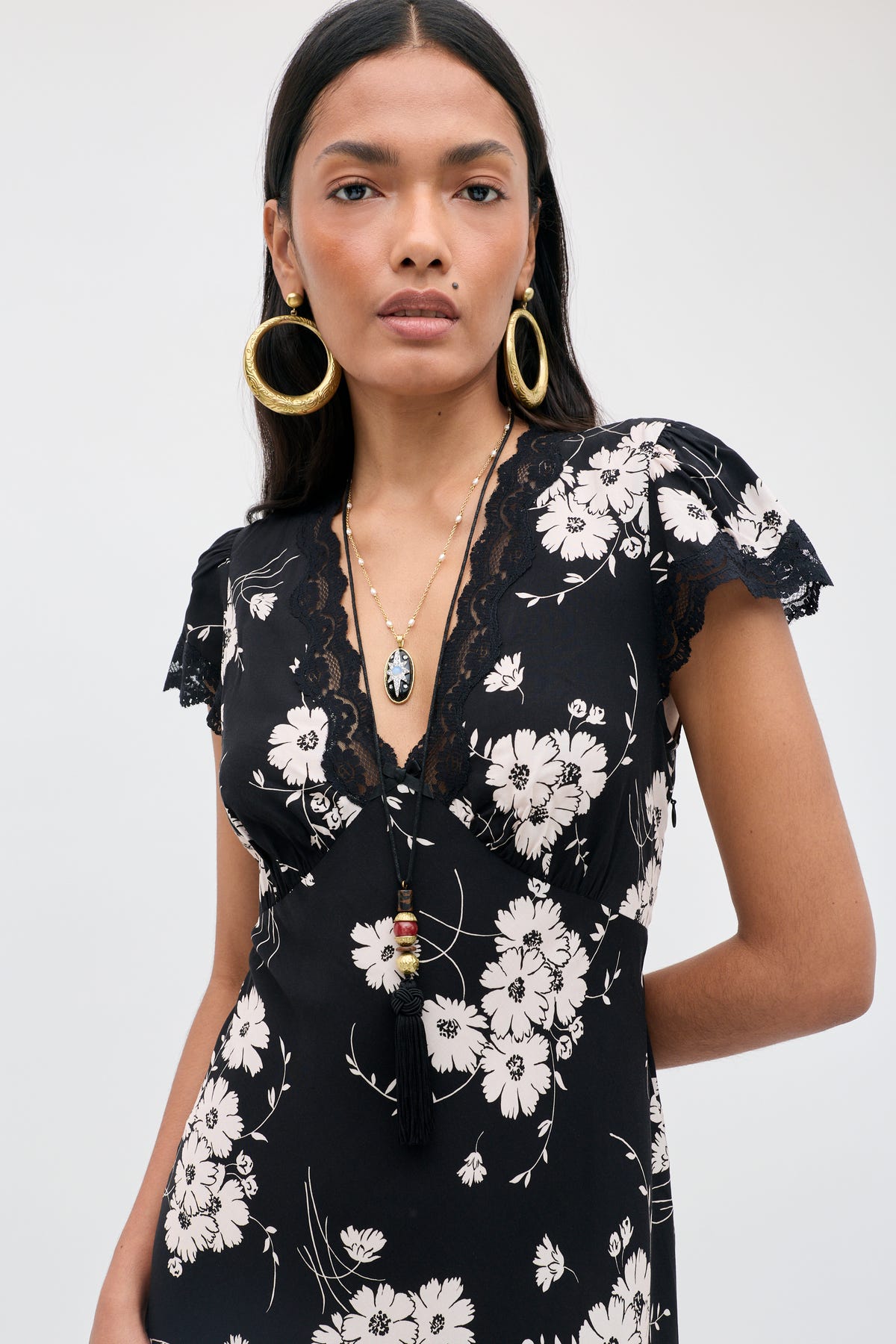 A woman wearing the Clarice - Daisy Field Black dress, which features a black base with a white daisy floral print and delicate lace trim on the sleeves and neckline. She accessorizes with large gold hoop earrings and layered necklaces, standing against a plain light background.
