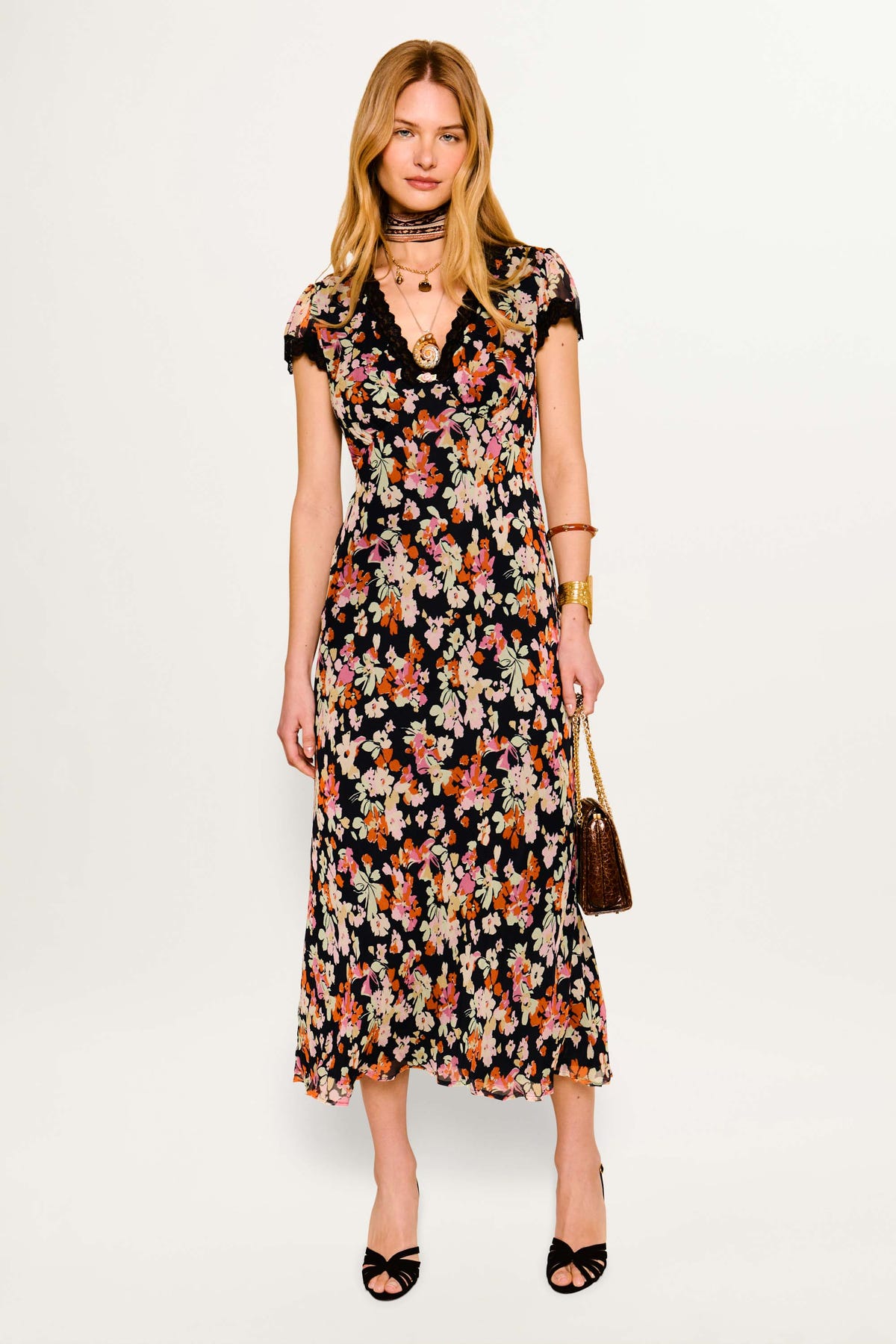 A woman stands against a white background wearing the Clarice - Primrose Floral Deep Navy dress, paired with black open-toe heels, layered necklaces, bracelets, and carrying a small textured handbag.