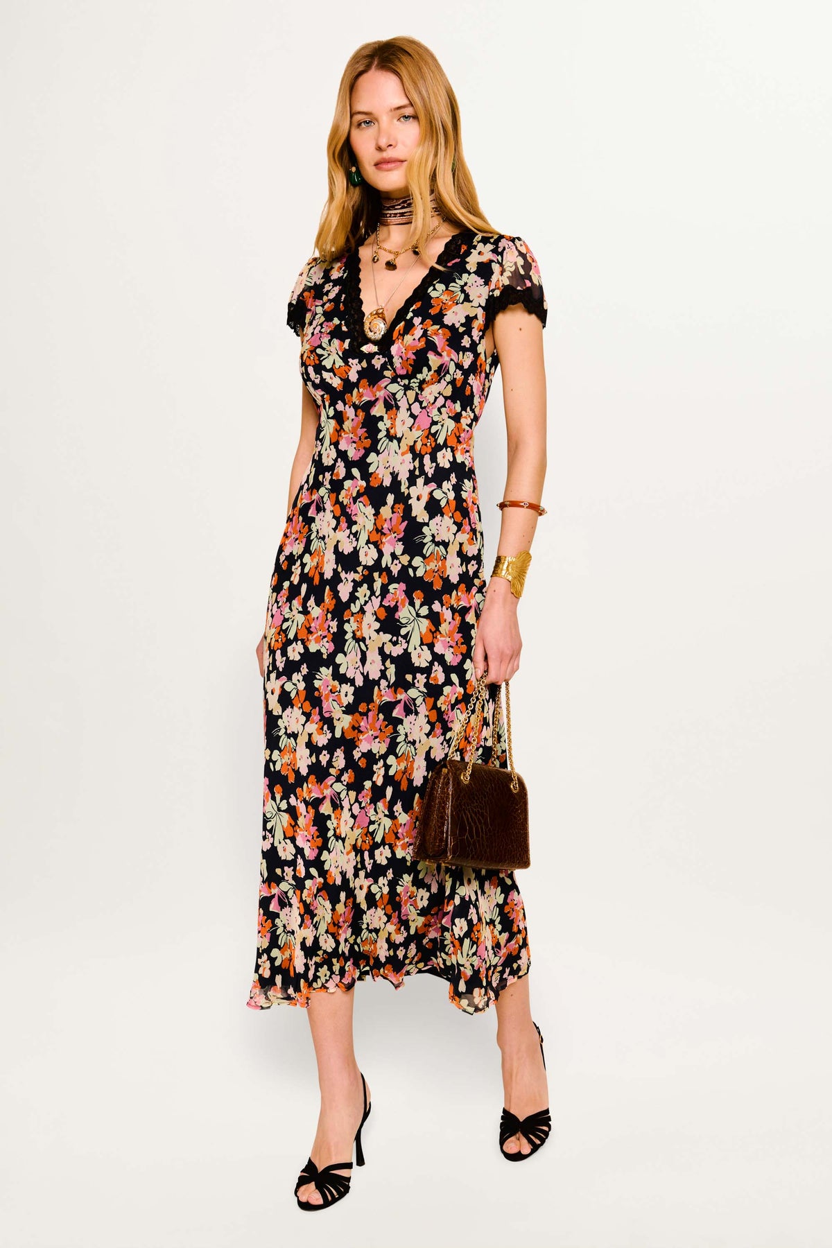 A woman wearing the Clarice - Primrose Floral Deep Navy dress, black heeled sandals, gold jewelry, and carrying a brown handbag stands against a white background—an elegant look for a day out.
