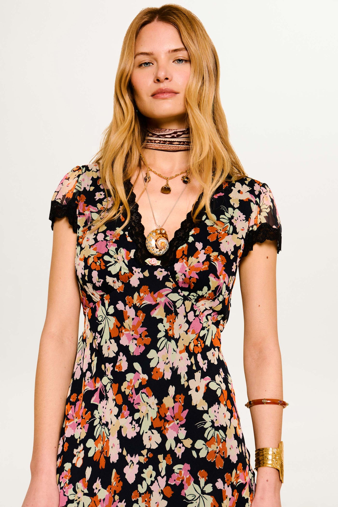 A woman with long blonde hair wears the Clarice - Primrose Floral Deep Navy dress, accessorized with layered necklaces, a patterned choker, and gold bracelets, standing against a plain light background.